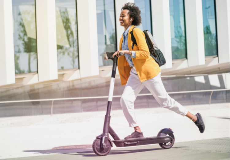 Junge Frau auf e-scooter mit gelben Blazer und weissen Hose.